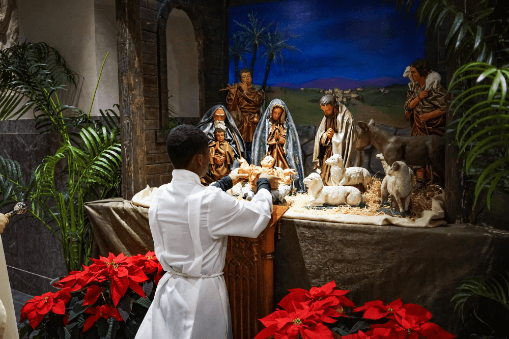 Altar server places a statue of the baby Jesus in the manger during the blessing of the nativity.