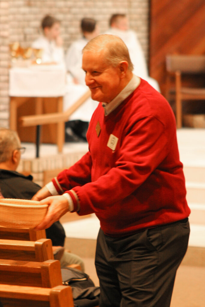 An older gentleman in a red sweater passes the offertory basket to parishioners.